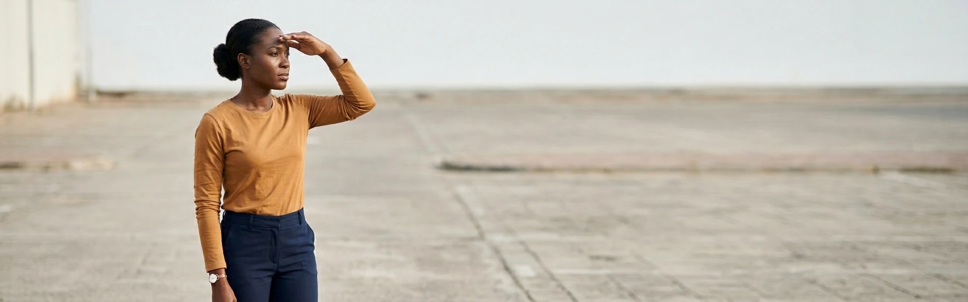 A Nigerian woman stands in an open paved area with one hand raised to shade her eyes as she looks into the distance