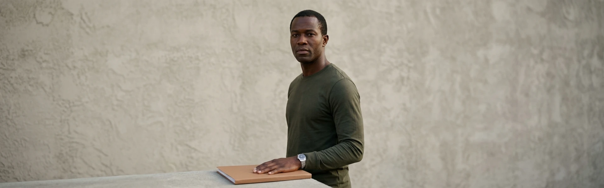 A Nigerian man stands with one hand resting flat on a plain closed folder on a low surface